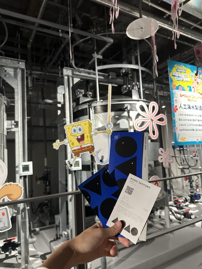 A person holding tickets in front of a modern industrial-style aquarium interior with a SpongeBob SquarePants character cutout in the background.